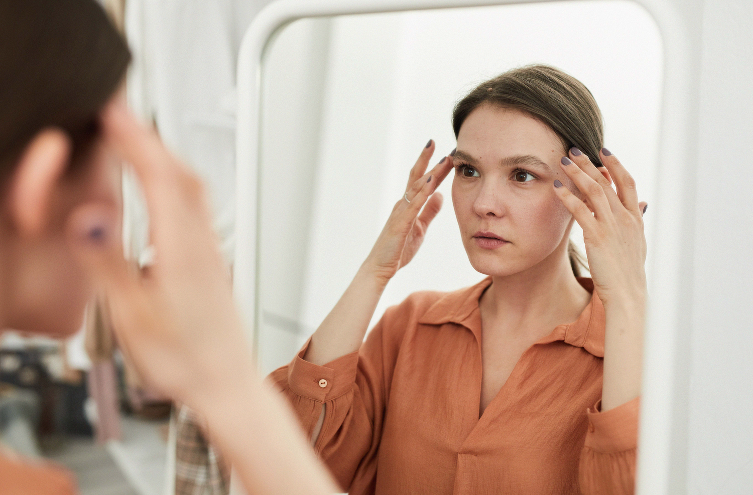 Woman looking into a mirror while gently touching her temples, wearing a rust-colored blouse in a softly lit, minimal interior.
