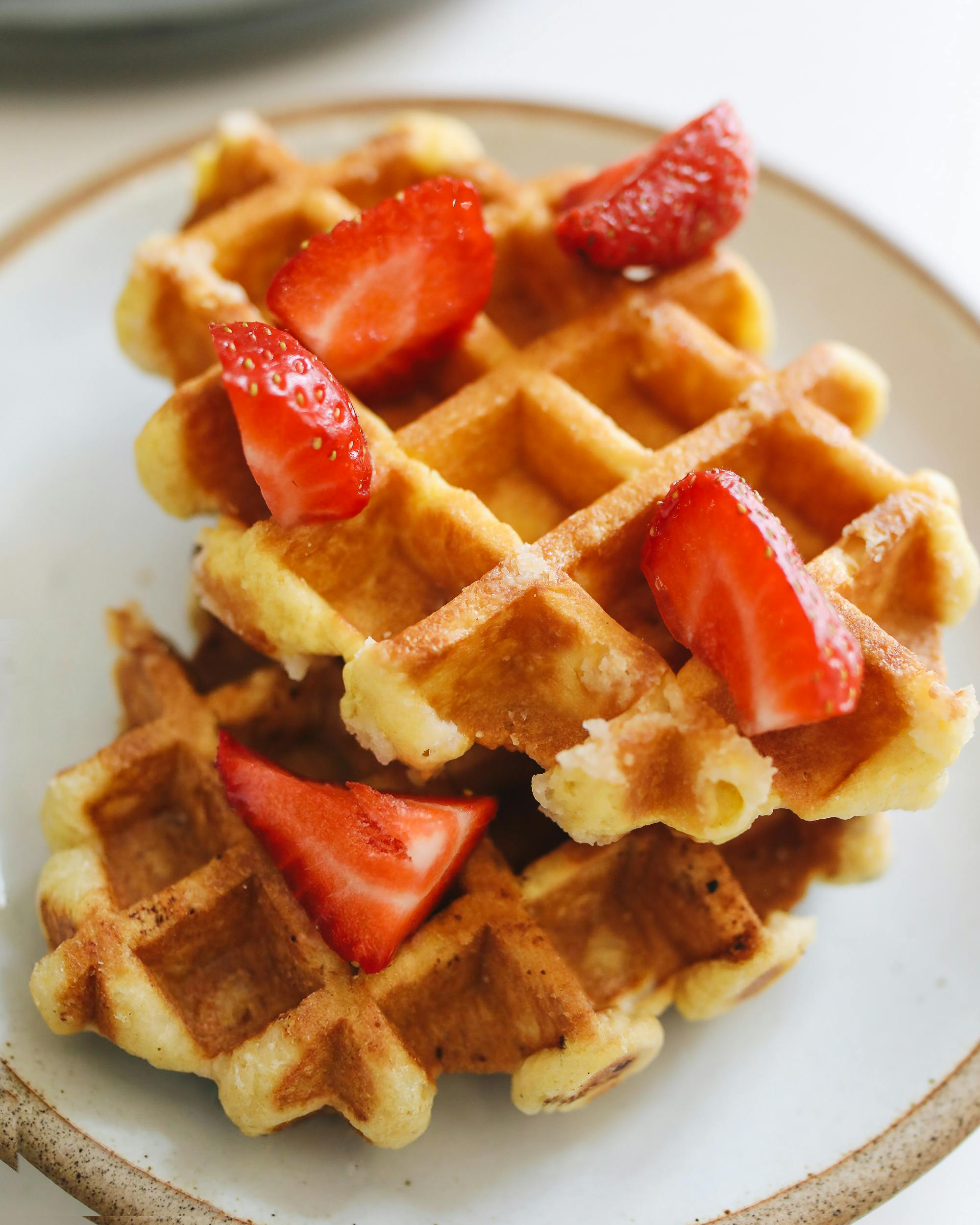 Stack of golden waffles topped with whole strawberries and served with sliced strawberries on a white plate.