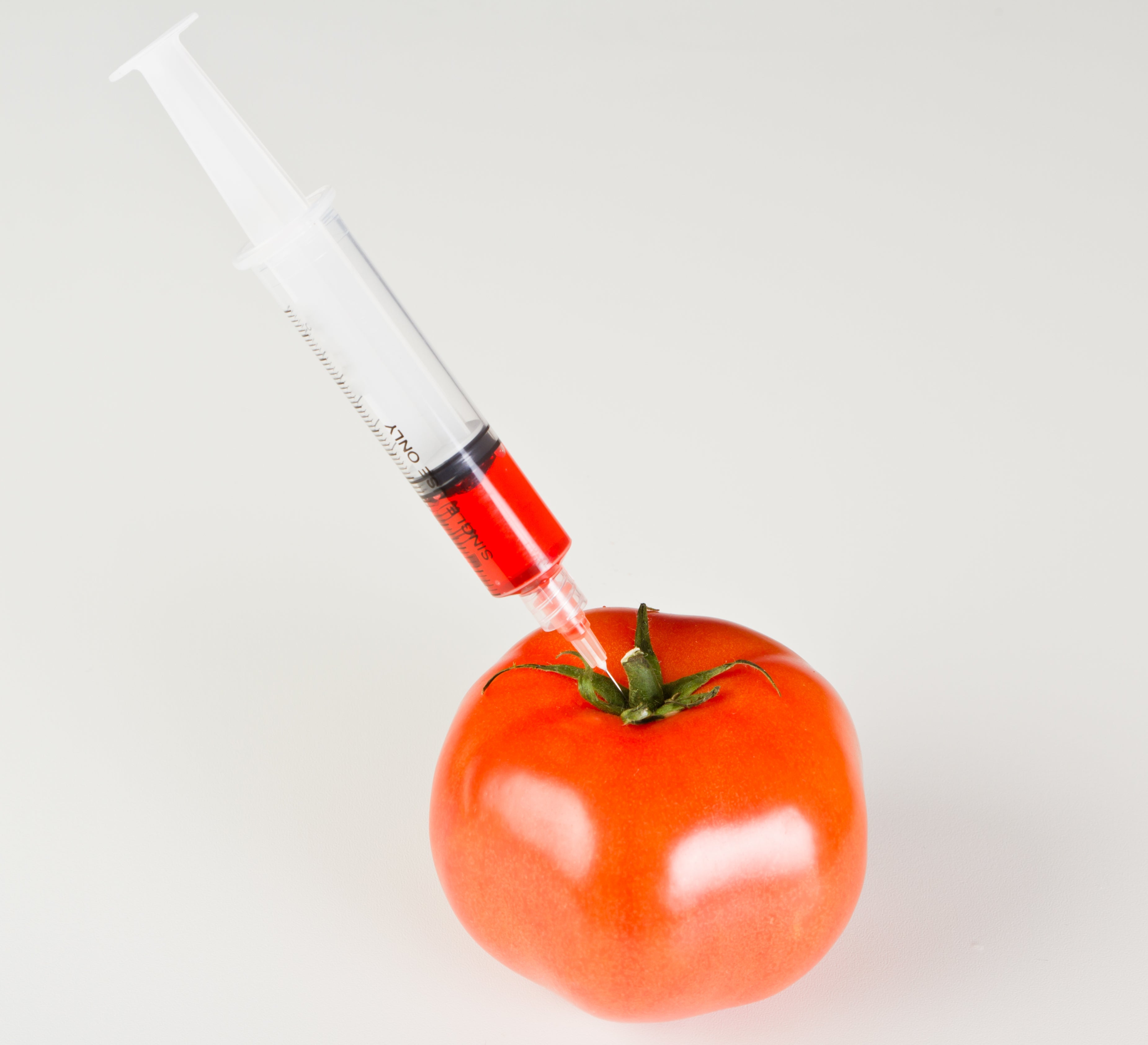 Syringe injecting a tomato with red liquid against a plain background.