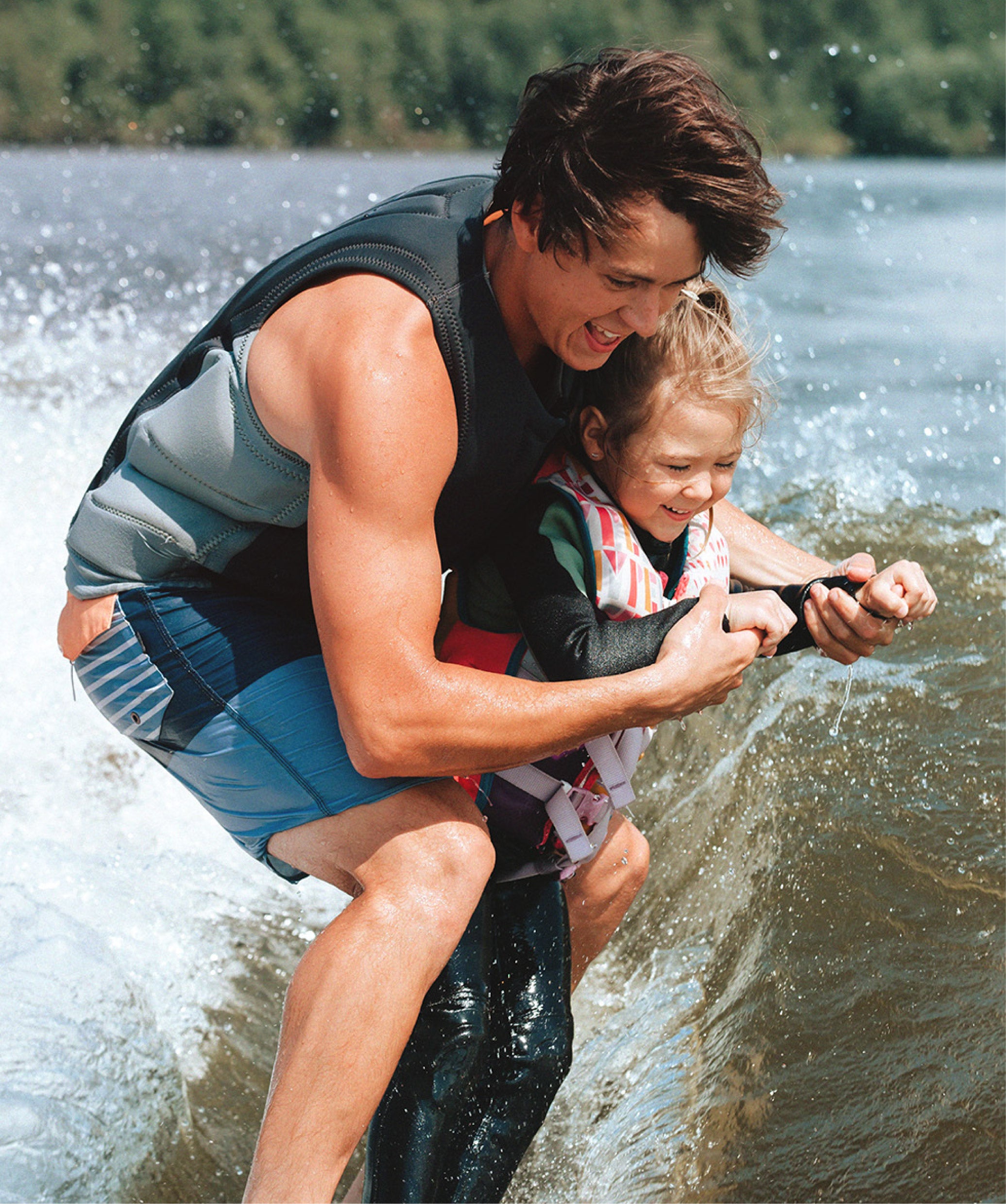 A father and a young child in water sports gear wakeboarding on a lake, with the man holding the child securely as they glide through splashing water.
