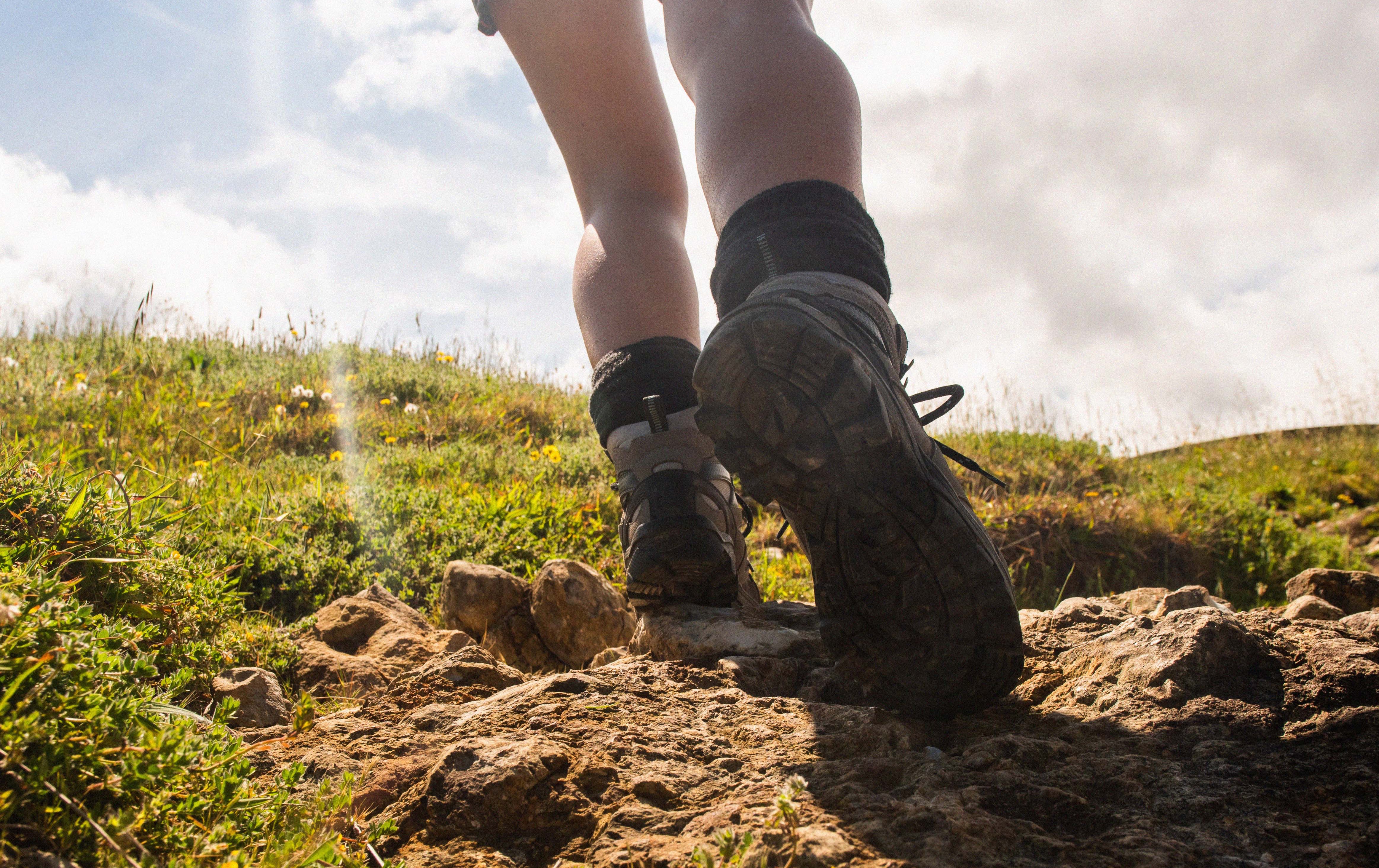 A low-angle outdoor shot of a person hiking up a rocky grassy trail, showing their legs, black athletic socks, and hiking shoes against a bright sky with clouds.